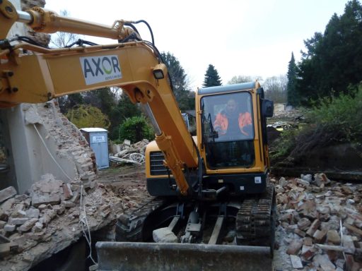 Bagger mit ausgefahrenem Arm, umgeben von Trümmern einer abgerissenen Struktur.