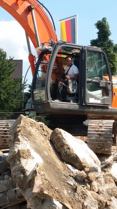 Baggerfahrer auf Baustelle mit deutscher Flagge im Hintergrund, große Steine daneben.